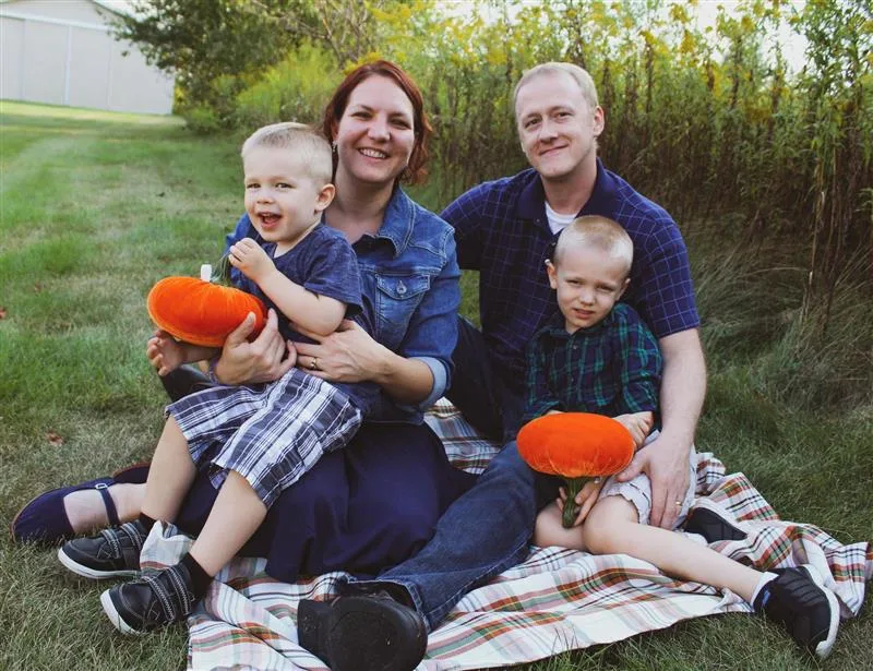 The image shows Jessica Wilbur, the ABS Kids Regional Recruiting Manager, with her family. They are seated together on a blanket outdoors, with two young children smiling and holding small orange pumpkins. Jessica, along with her partner, is enjoying a happy family moment, with their arms around their children. The setting looks like a pleasant outdoor fall scene.