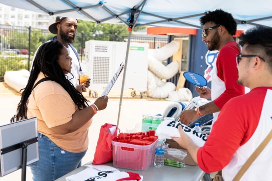 A group of ABS Kids staff members are pictured at an event booth, engaging with community members. One staff member hands out branded materials, while others stand by, ready to assist. The booth is filled with ABS Kids merchandise such as T-shirts and promotional items. The image reflects the organization's commitment to supporting families and individuals in the autism community through outreach and engagement.