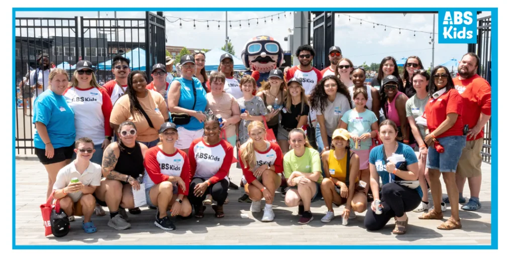 The image captures a group photo of ABS Kids staff and community members gathered at a sensory-friendly event. The group, all wearing ABS Kids branded shirts, stands in front of a welcoming entrance with colorful decorations. The event highlights ABS Kids' commitment to providing inclusive and sensory-friendly experiences for individuals with autism, reflecting their dedication to supporting the community.