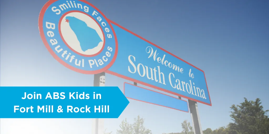 Alt text: "Welcome sign to South Carolina with the text 'Smiling Faces, Beautiful Places,' and a banner below reading 'Join ABS Kids in Fort Mill & Rock Hill,' promoting career opportunities with ABS Kids in South Carolina.