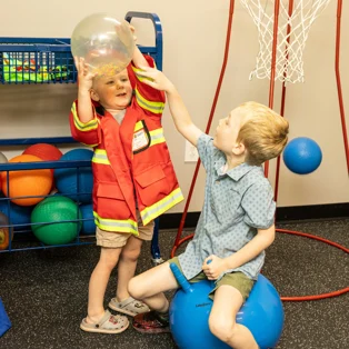 Two young children playing in a therapy room at ABS Kids. One child is wearing a red fireman jacket and holding a large clear ball while the other child is sitting on an exercise ball, reaching up to assist. Colorful therapy balls are visible in the background along with a basketball hoop.