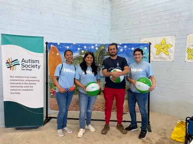 A group of four people standing together at an event, holding beach balls with the ABS Kids logo. They are standing in front of a backdrop with a starfish decoration and a banner for the Autism Society San Diego. The group is wearing ABS Kids t-shirts, and the banner behind them reads: 'Our mission is to create connections, empowering everyone in the autism community with the resource needed to live fully.' Keywords: ABS Kids, Autism Society San Diego, beach balls, autism, event.