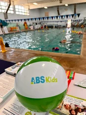 A beach ball with the ABS Kids logo in the foreground, placed on a table at a swimming pool. The pool is visible in the background with swimmers and poolside activities. Keywords: ABS Kids, swimming, beach ball, poolside.