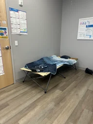 A folding cot with a blanket and pillow on it, placed in a room with light gray walls and hardwood floors. A door with a white frame is visible, as well as a few posted documents on the wall. The setting suggests a quiet or resting area within an ABS Kids facility.