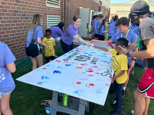 Group of children and volunteers participating in an outdoor painting activity. Kids are stamping their handprints on a large white canvas, while volunteers assist them. They are wearing matching purple t-shirts and yellow shirts. Keywords: ABS Kids, outdoor activity, handprint painting.