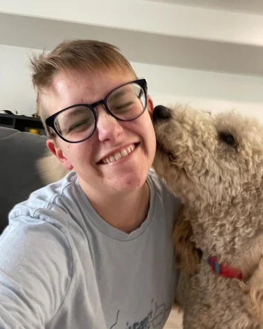 This image features Savannah Sivertson, a person with short hair and glasses, smiling as a goldendoodle affectionately licks her cheek. Savannah is wearing a light-colored t-shirt and appears joyful in this candid moment. The background is a neutral interior setting, suggesting a cozy home environment.