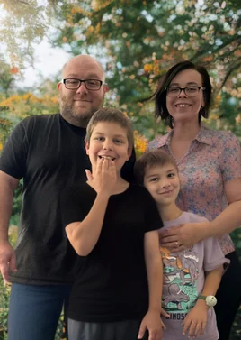 A family photo featuring Robin Masciave, an autism mom and advocate, standing with her partner and two children. The family is smiling in a natural outdoor setting with trees in the background. The children are standing in front of their parents, one boy is smiling and the other is playfully covering his mouth with his hand.