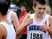 A young male athlete, wearing a white running vest with the number 1988, gives two thumbs up while standing in a crowd of other runners at a cross-country event. He is smiling confidently as he prepares for the race.