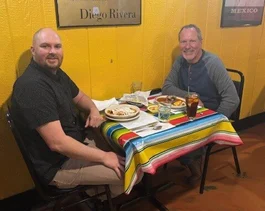 Two men sitting at a table in a restaurant, smiling and enjoying lunch. The table is covered with a colorful striped tablecloth, and each person has a plate of food in front of them. One man is wearing a black shirt, and the other is in a gray long-sleeve shirt. A glass of drink is visible on the table.