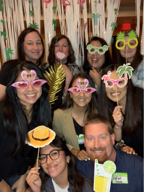 Group photo of ABS Kids team members posing with fun tropical-themed props like flamingo glasses, sunglasses, and hats at a festive event, smiling and having fun.