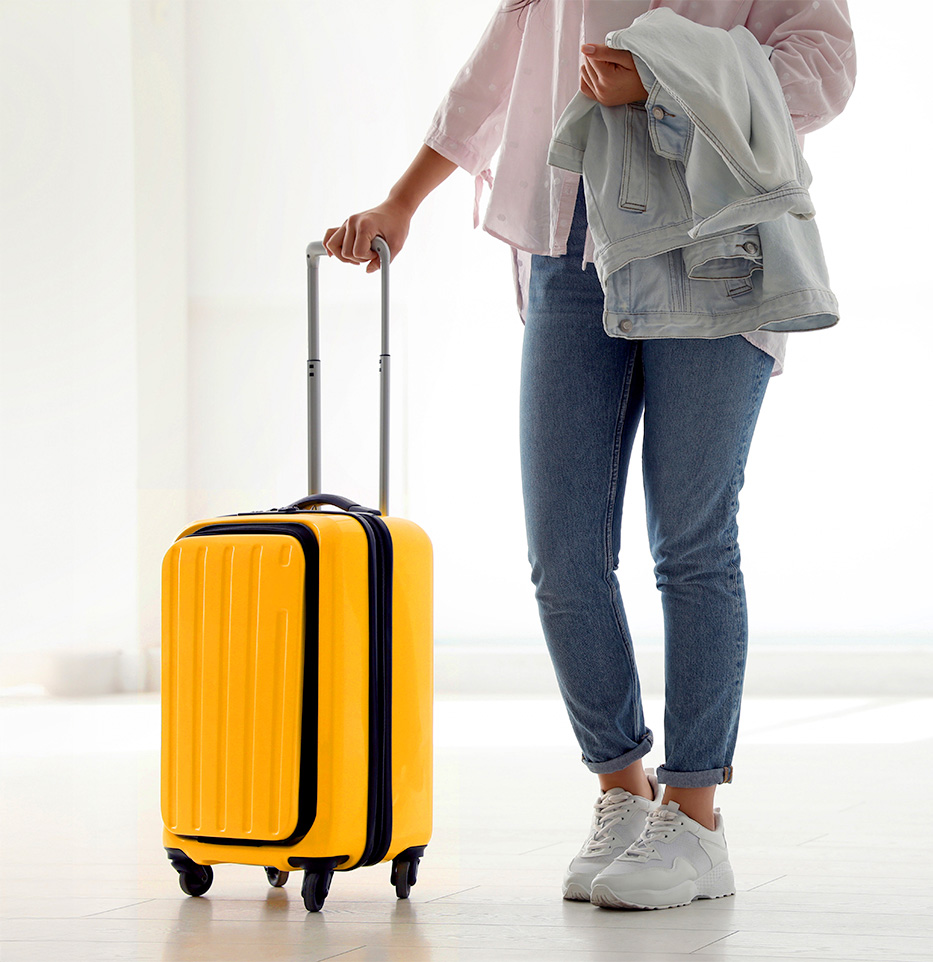 A bright, sunlit interior shot showing the lower half of a woman standing next to a vibrant yellow carry-on suitcase. She’s wearing rolled-cuff blue jeans and white sneakers, holding a folded light denim jacket in her left arm while her right hand grips the suitcase’s extended telescoping handle.